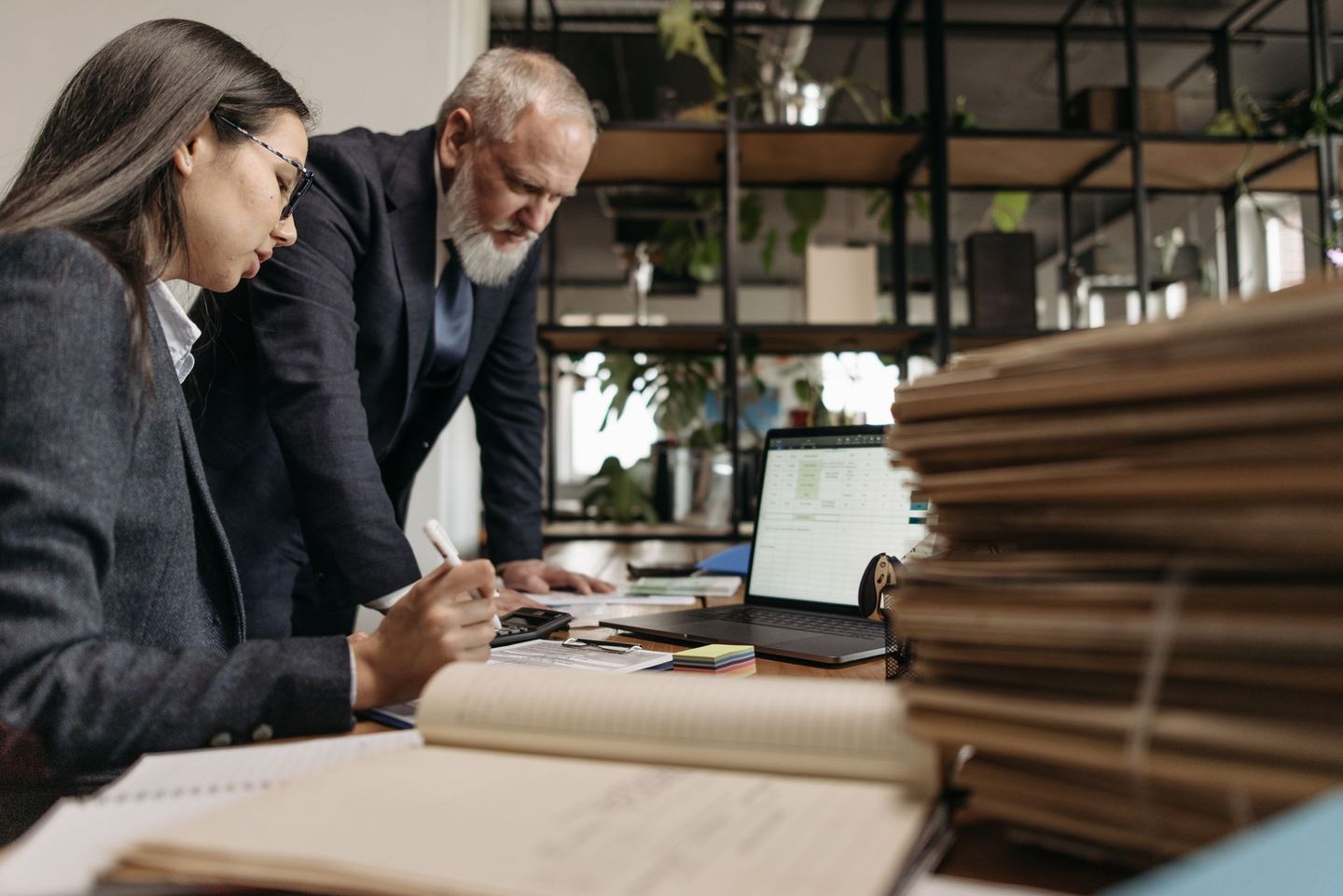 Two colleagues at a desk going down a spreadsheet on a laptop beside a stack of files